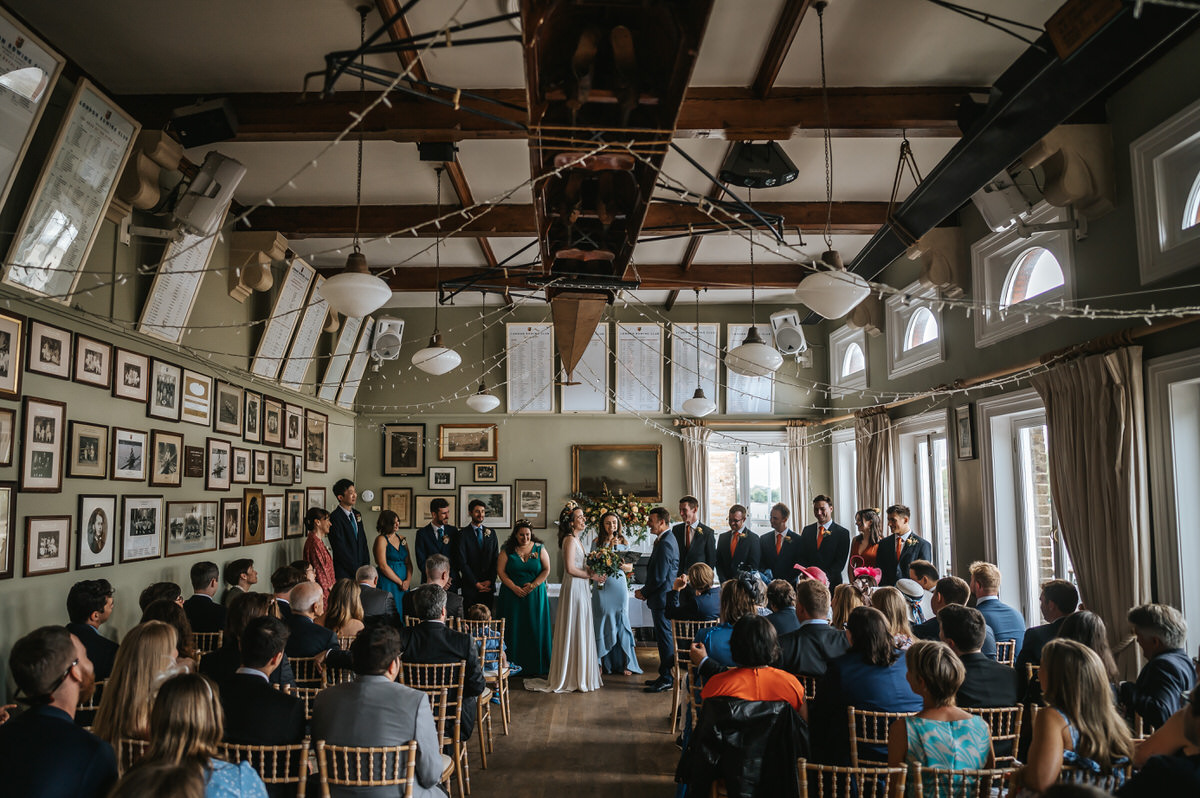 london rowing club wedding ceremony showing the whole room from the back with the newlyweds in the middle and guests to the sides