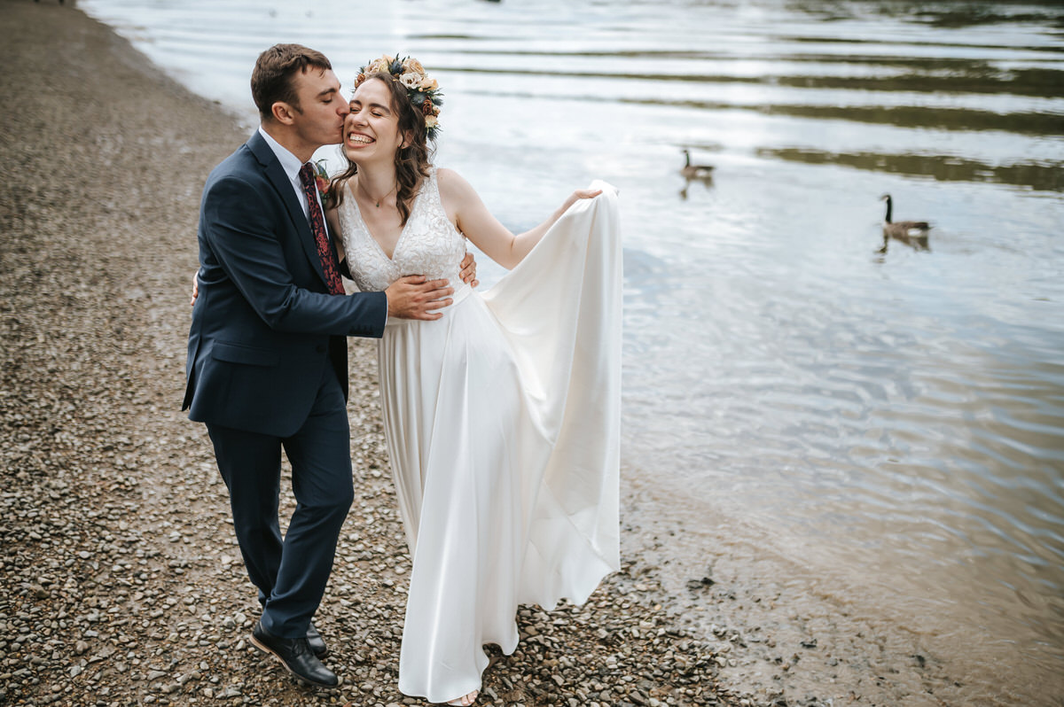 london rowing club wedding groom kissing the bride on her cheek as they walk along river thames