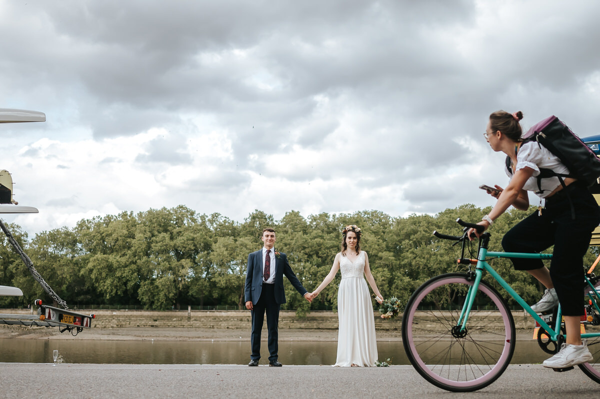 london rowing club wedding couple holding hands with river thames behind them and a person on a bicycle looking at them as they ride by